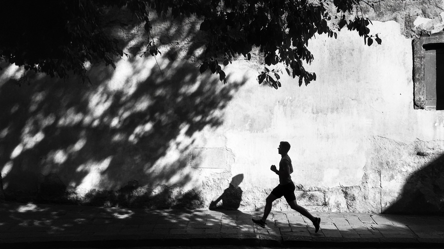 Silhouette of a man jogging along a sidewalk, with shadows of trees cast on a textured wall in the background